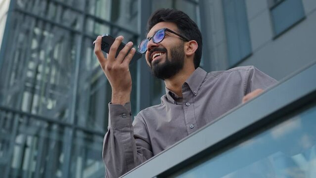 Happy smiling Indian man businessman business employer guy record voice message on phone audio chat dictate voicemail in microphone using AI mobile assistance speak smartphone talk loudspeaker in city