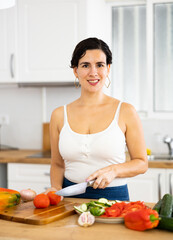 Cheerful young Hispanic woman following healthy diet preparing vegetable salad for dinner in home kitchen