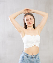 Fototapeta premium Teenage girl in white strapless top, jeans stands and holds hands above head, joining palms together. Advertising of cosmetics, care procedures, isolated on gray background