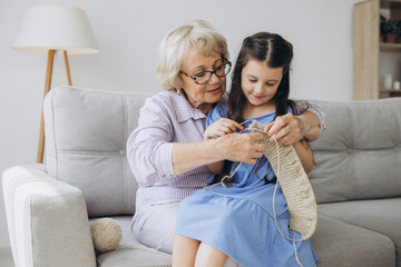 Grandmother teaching her pretty little granddaughter to knit as they sit close together on a sofa in the living room