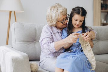 Grandmother teaching her pretty little granddaughter to knit as they sit close together on a sofa in the living room