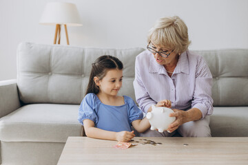 Obraz na płótnie Canvas Grandmother and her granddaughter Putting Coin Money In Piggybank At Home. Personal Savings, Bank Safety And Financial Investments Concept