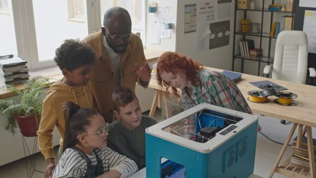 High angle shot of kids looking how 3D printer works while teacher talking to them during computer class in school