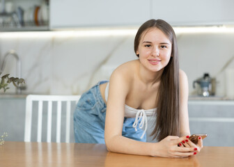 Young girl in casual attire put elbows on kitchen table, props chin with fist and holds smartphone...