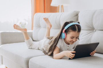 Happy little girl sitting on sofa, wearing headphones and using tablet. Technology, childhood and domestic life.