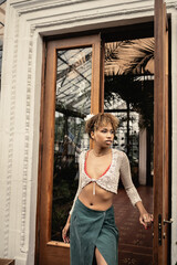 Confident young african american woman in knitted top and summer outfit looking away while standing near open door of indoor garden, fashionable woman enjoying summer vibes