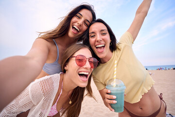 Three excited young Caucasian friends in summer clothes taking selfie on beach. Group of smiling women enjoying vacation. Cheerful beautiful girls of generation z pose for photo with mobile phone. - Powered by Adobe