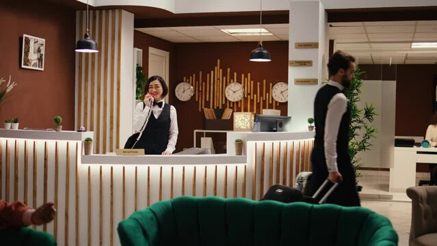 Happy professional hotel personnel doing various administrative tasks in stylish hotel reception. Asian receptionist taking room booking phone calls and bellboy carrying tourist luggage