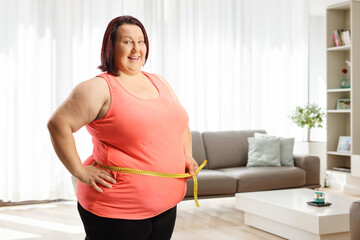 Overweight woman measuring her waist with a tape and smiling in a living room