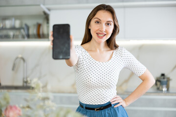 Smiling woman student showing modern smartphone with blank screen in kitchen