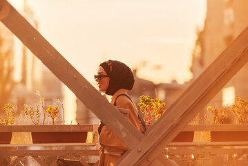A hijab woman in stylish sunglasses and an elegant French outfit, walking through the city at sunset, carrying a bouquet, bread, and newspaper, radiating a sense of cultural charm and serenity