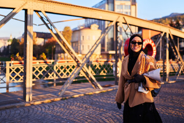 A hijab woman in stylish sunglasses and an elegant French outfit, walking through the city at sunset, carrying a bouquet, bread, and newspaper, radiating a sense of cultural charm and serenity