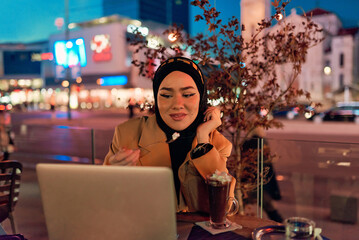 A hijab woman enjoying a sweet dessert at a coffe while working on her laptop during the nighttime...