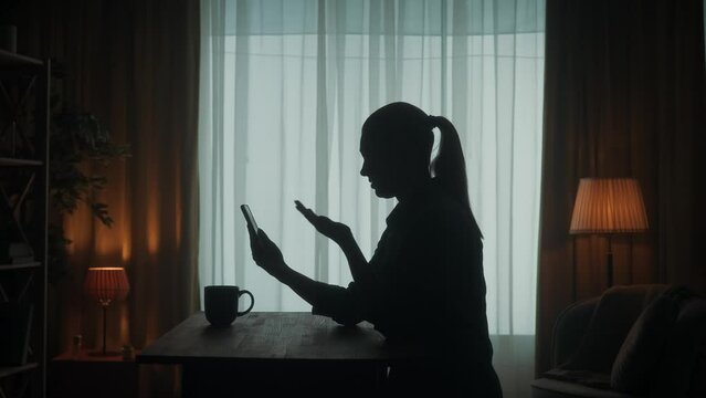 A woman is talking on a video call on the phone while standing at a table in the living room. A woman waves her hand in greeting, talks, says goodbye. Side view of the dark silhouette of a woman.