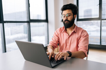 Fototapeta premium Focused Indian freelancer male in stylish eyeglasses working on laptop sitting at desk in light coworking office room by window. Bearded business man wearing casual clothes looking at computer screen.
