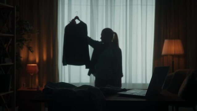 A woman trying to choose what to wear. A woman holds two blouses on a hanger, shrugs her shoulders, can't decide which one to wear. Dark silhouette of a woman in the living room.