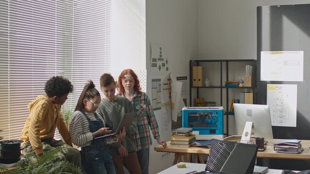 Group of multi-ethnic kids standing by window in school classroom and discussing programming code on laptop during computer class