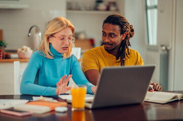 Multicultural spouses are sitting in their cozy home and doing home finances on the laptop online.