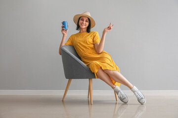 Beautiful woman with can of soda sitting in soft armchair near grey wall