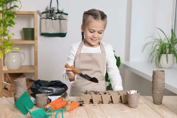 Little girl adding soil into peat pots at wooden table in room. Growing vegetable seeds