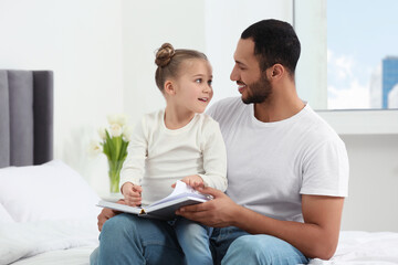 Fototapeta premium Little girl with her father reading book together on bed at home. International family