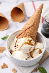 Scoops of ice cream with caramel sauce and wafer cone on white marble table, closeup