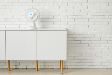 Stylish cabinet with modern electric fan near white brick wall