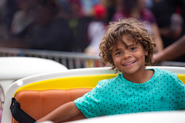 Cute diverse little boy smiling while riding an amusement park carnival ride outdoors. Fun summer activity. A beautiful expression of joy