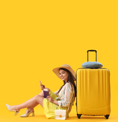 Young woman with passport and suitcase using mobile phone on yellow background