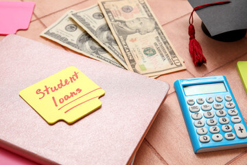Sticky note with text STUDENT LOANS, dollar banknotes, calculator, notebook and graduation cap on tiled background