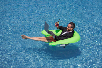 Freelance work, distance online work, e-working. Summer business. Business man in suit drink summer cocktail and using laptop in pool. Businessman dreams on summer business in swimming pool water.