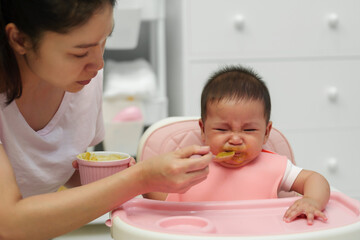 mother feeding food to her crying infant baby with spoon, refuse to eat