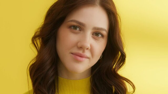 Pretty woman with dark styled curls, wearing yellow sweater, posing on yellow background. Medium close up. Woman looks down, bites lips, feeling shy. High quality 4k footage