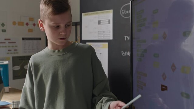 Boy explaining computer algorithm scheme on digital screen while giving presentation during programming lesson at school, black teacher listening in background
