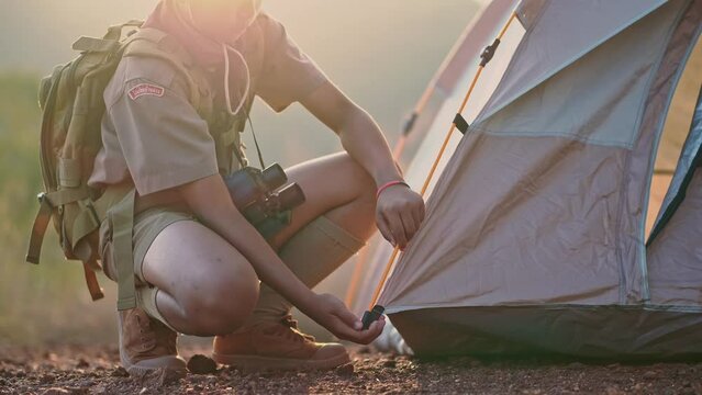 An Asian Boy Scout Sits on the Ground and Packs his Backpack in a Mountain Scout Camp near Sunset.