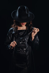Fototapeta na wymiar Sexy female dominant mistress with a whip for BDSM sex with submission and domination in a leather raincoat and a hat in the noir style