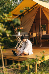 Young woman works on laptop while sitting relaxed on chair by the campfire, traveling with tent. Concept of remote work and escape to nature