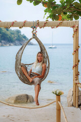 Relaxed caucasian tanned young woman with blonde hair sitting on a wicker swing on the beach. Caucasian girl in relaxed pose sits on swing during her summer vacation on tropical beach. Happy tourist