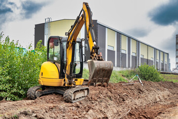 Yellow mini excavator at the construction site on the edge of a pit against a cloudy blue sky....