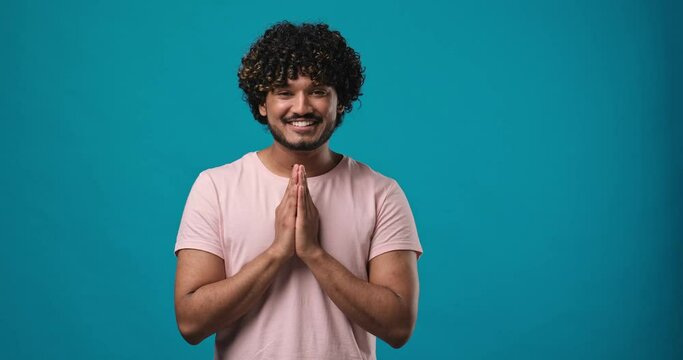 Young bearded indian man making Namaste greeting gesture with slight bow standing isolated on blue studio background