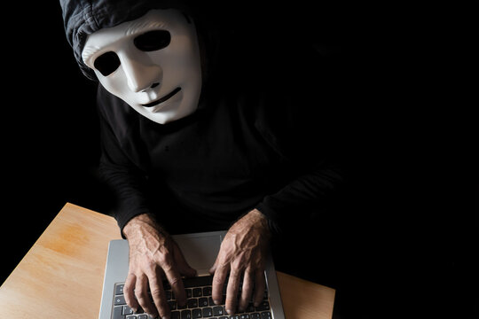 hacker dressed in black with mask and hood typing on a computer while looking at the camera cybercrime concept