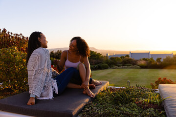 Biracial lesbian couple sitting on mattress in garden at sunset, copy space