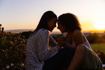 Biracial lesbian couple sitting and holding hands in garden at sunset, copy space