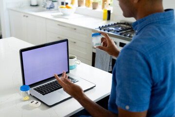 Fototapeta na wymiar Biracial man holding medication making video call in kitchen using laptop with copy space on screen