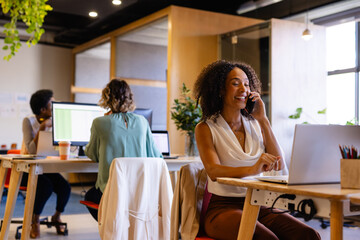 Obraz premium Happy biracial casual businesswoman using laptop and talking on smartphone in office