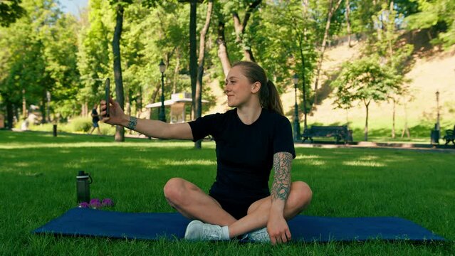 Portrait of a young female athlete on a sports mat resting after a workout in the park taking a selfie on a smartphone the concept of a healthy lifestyle