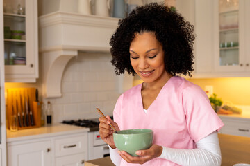 Happy biracial female medical worker in scrubs eating bowl of breakfast cereal in kitchen at home