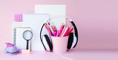 On the table on a pink background are pink school supplies, pencils, felt-tip pens, white sheets of paper, notebooks, a notepad and headphones.  Concept back to school.  Space for copy text.