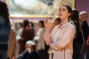 Portrait of young Caucasian woman holds glasses and looking at exhibition. People and masterpieces in background. Concept of Museum Day