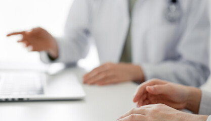 Doctor and patient sitting at the desk in clinic office. The focus is on female physician's hands pointing to laptop computer monitor, close up. Perfect medical service and medicine concept